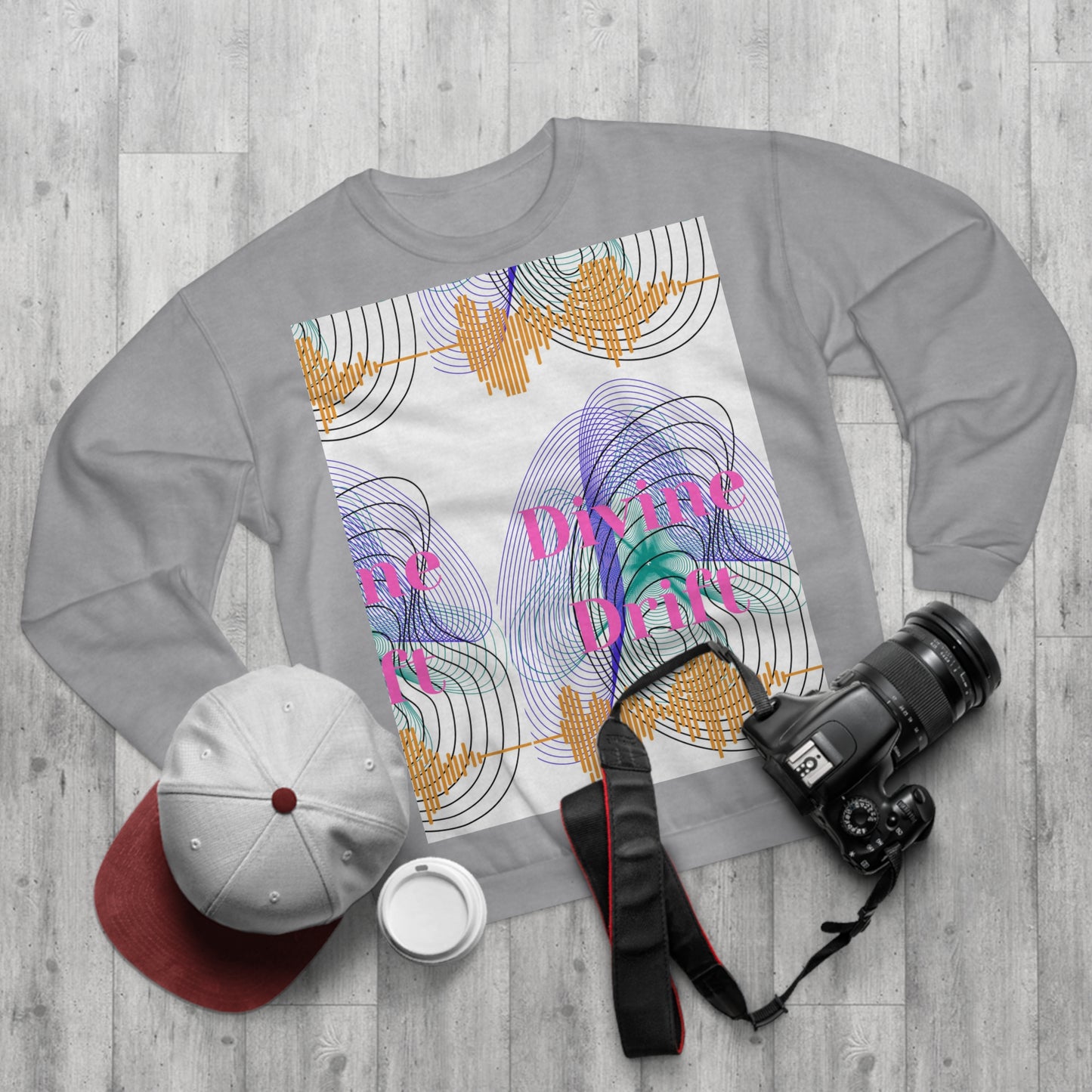 Gray sweatshirt with colorful design and text on a wooden surface with a cap, camera, and cup.