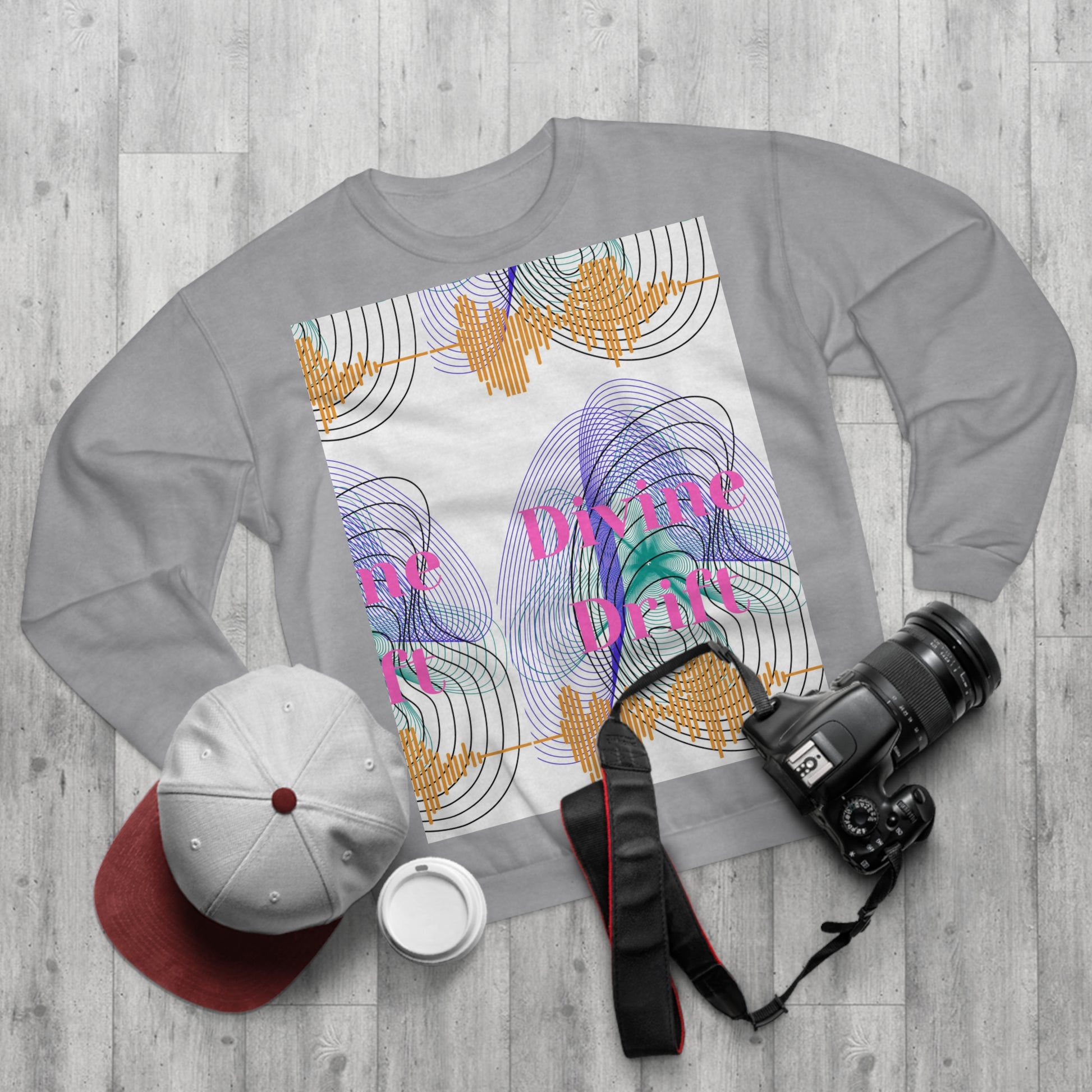 Gray sweatshirt with colorful design and text on a wooden surface with a cap, camera, and cup.