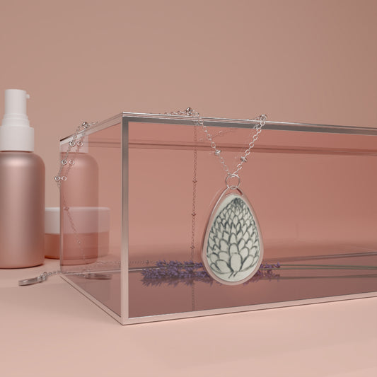 Teardrop-shaped pendant with floral design on a chain, placed inside a clear box with skincare products in the background.